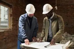 Two biracial builders with helmets on heads having discussion about renovation while concentrated on layout